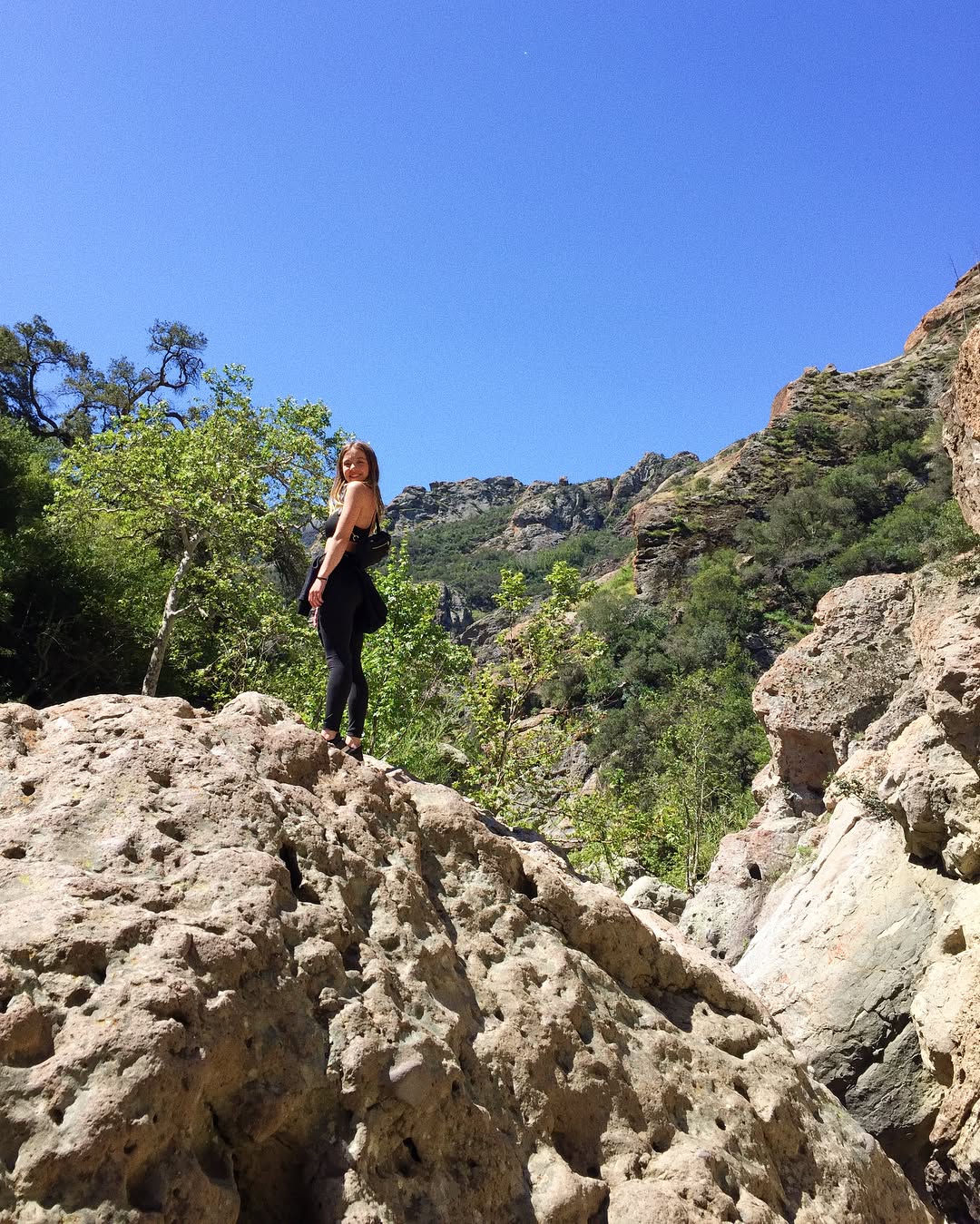 a young woman with a backpack stands on a large rock formation, looking back towards a stunning view of a rocky canyon under a bright blue sky. the landscape is arid and mountainous, with sparse vegetation. the woman is dressed in black athletic wear, suggesting she's hiking. the image captures a sense of adventure and exploration in a beautiful natural setting.