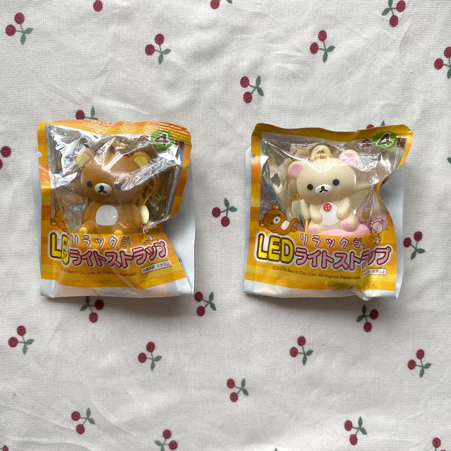 the image shows two rilakkuma led light straps in their original packaging, lying on a surface with a cherry pattern. one is a brown rilakkuma and the other is a beige korilakkuma. the packaging has japanese text and logos.