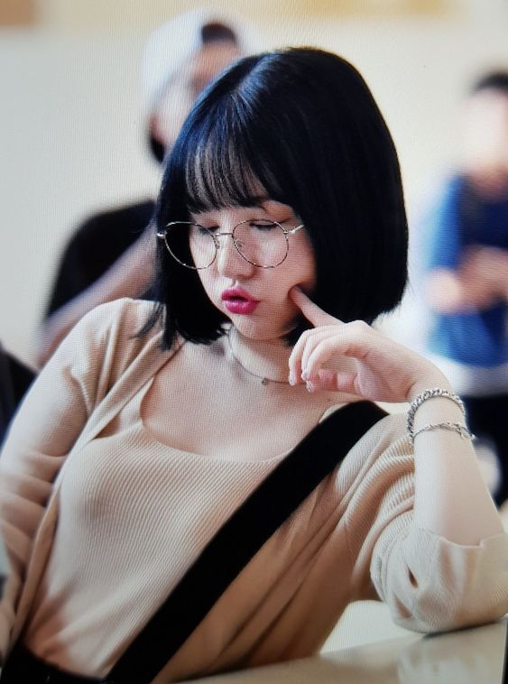 a young woman with a black bob haircut and glasses sits leaning against a surface. she is wearing a beige cardigan over a matching tank top, accessorized with a delicate necklace and bracelet. she has a soft, dreamy expression, with her cheek resting on her hand and her lips slightly parted. the lighting is soft and natural, highlighting her youthful features.  the overall aesthetic suggests a korean fashion influence.