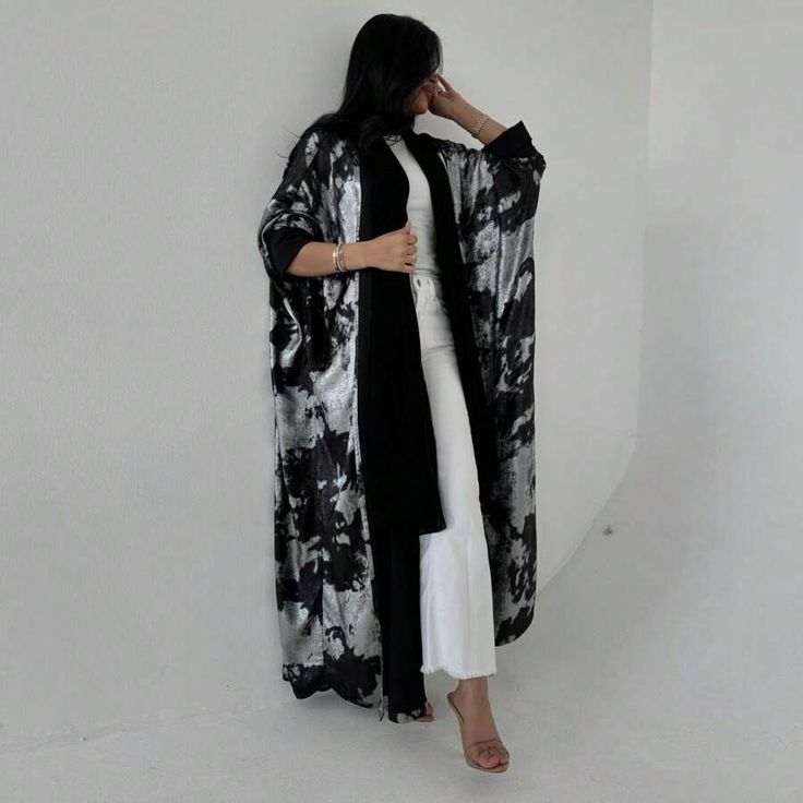 a woman is standing against a plain white wall, posing in a stylish black and silver abaya. she is wearing a white top and white jeans underneath the abaya. she has on a pair of high heels. she has several bracelets on her wrists and is touching her head with her hand. the background is minimalist, emphasizing the abaya.
