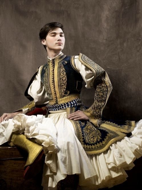 the image is a striking studio portrait of a young man elegantly dressed in a traditional greek fustanella costume.  he is seated, his posture poised and his expression serious. the costume is richly detailed; a dark blue velvet vest heavily embroidered with gold thread is worn over a white, ruffled fustanella (a pleated skirt).  the sleeves are long and similarly embroidered. the backdrop is a muted dark brown, which contrasts with the vibrant colors and textures of the clothing, making the young man the clear focal point. the overall feel is one of historical elegance and cultural significance.
