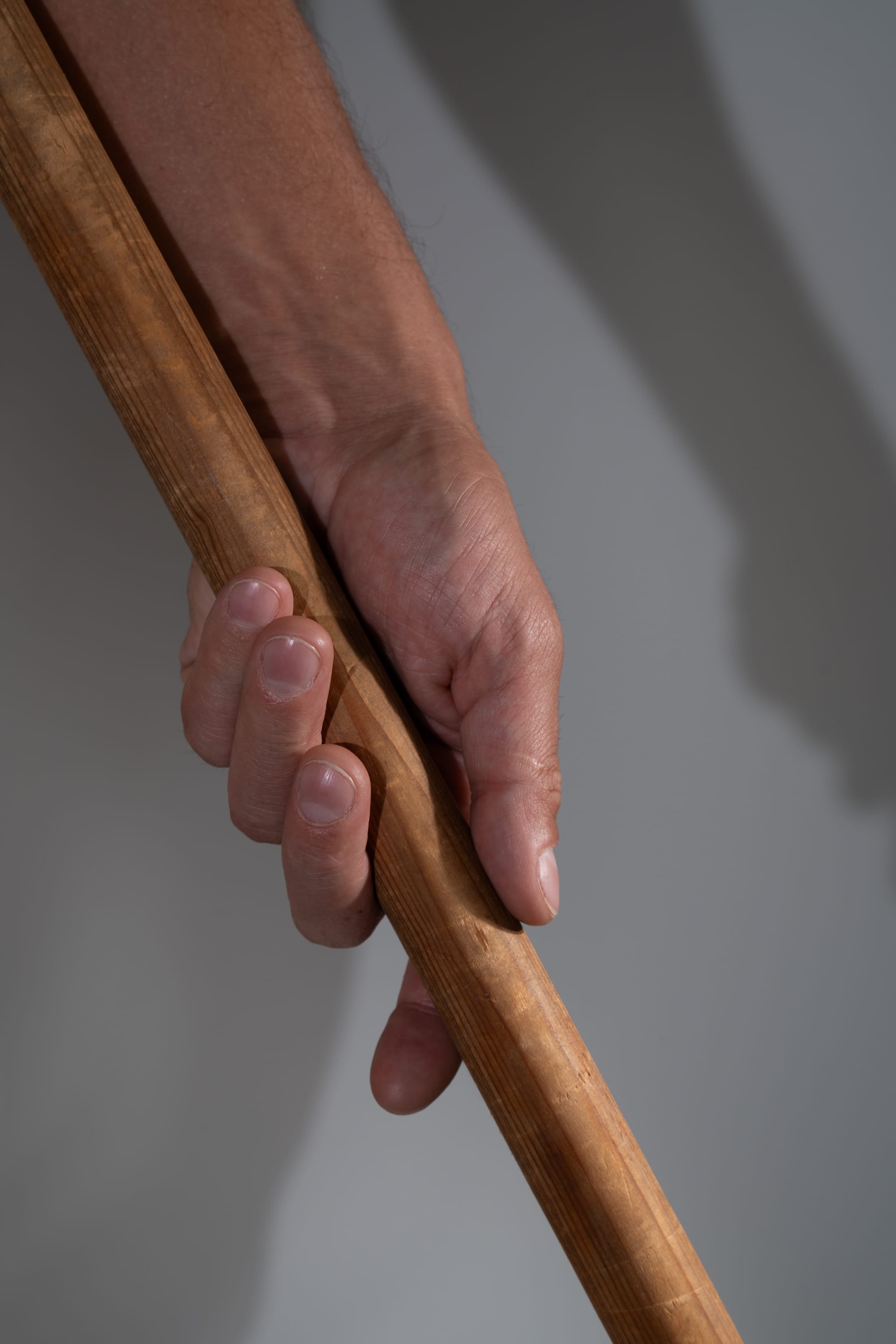 a closeup image of a man's hand tightly gripping a thick wooden stick or pole. the hand is illuminated by studio lighting, casting a distinct shadow on the background. the image emphasizes the strength and firmness of the grip.