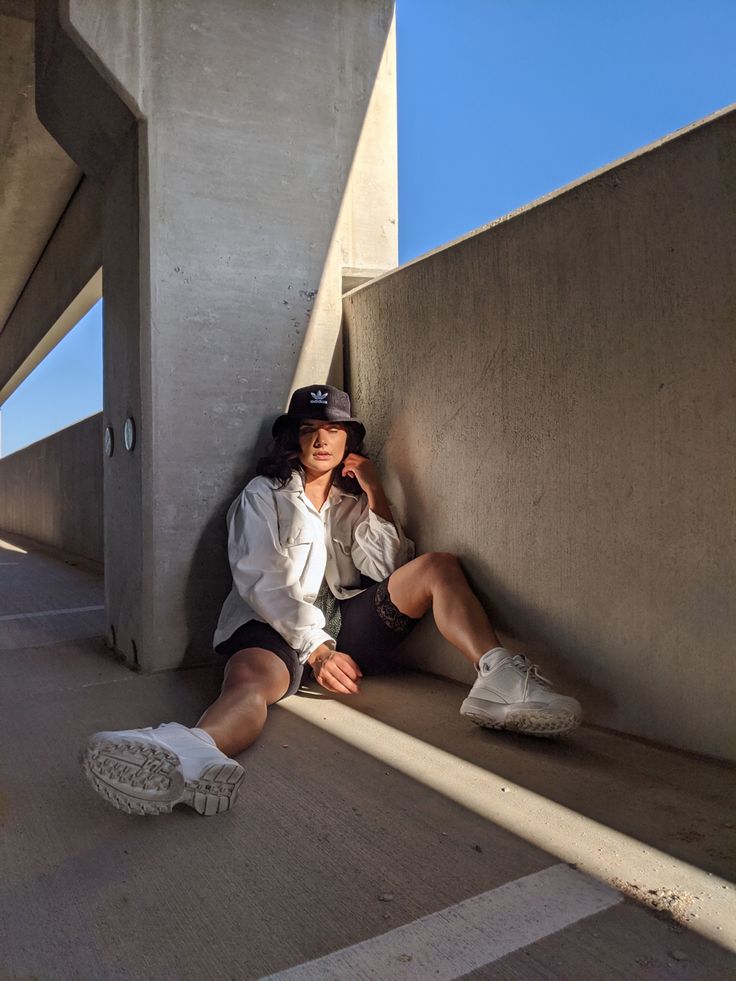 a young woman sits against a concrete wall in an urban setting. she is wearing a white shirt, black shorts, white sneakers, a black bucket hat, and appears to be posing for a photograph. the scene is bathed in sunlight, casting shadows on the concrete. the overall aesthetic is casual and stylish.
