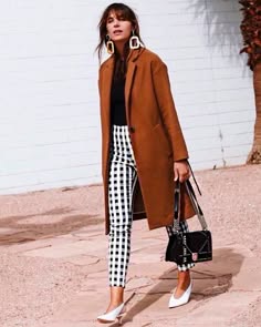 a woman is standing on the sidewalk, wearing a long brown coat, black and white checkered pants, white shoes, and a black handbag. she is also wearing large white earrings.