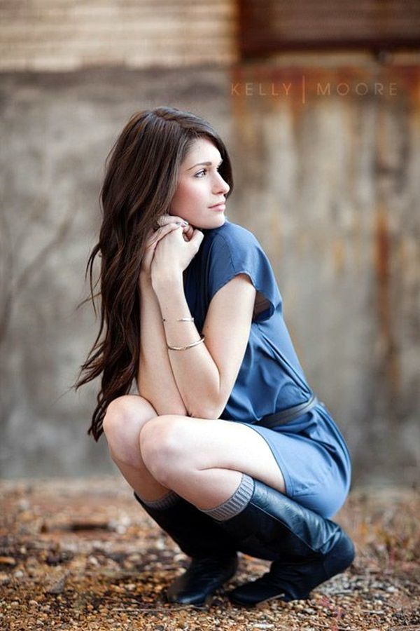 a young woman with long brown hair is crouching on a gravel surface, her back leaning against a weathered brick wall. she is wearing a blue tshirt dress and black boots, with her arms resting on her knees and her gaze directed thoughtfully off to the side. the setting appears to be an abandoned or industrial area, with muted, natural light illuminating the scene.  the overall atmosphere is one of contemplation and quietude.