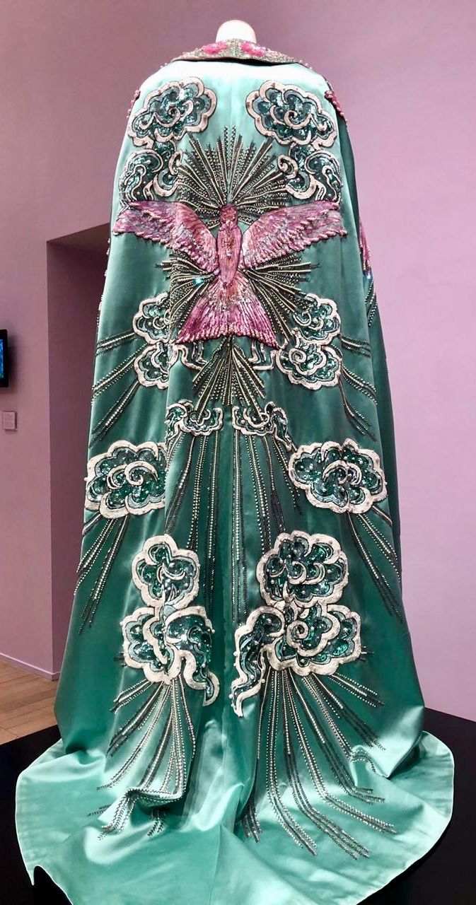 a turquoise green cape with elaborate embroidery on a mannequin. the design features a large pink bird with outstretched wings at the upper part of the cape, surrounded by stylized clouds that are outlined in white and filled with green sequins. below the bird are additional cloud motifs and radiating lines made of silver beads or sequins. the cape is displayed on a black platform with a pink wall in the background, suggesting it is a museum or gallery exhibit.