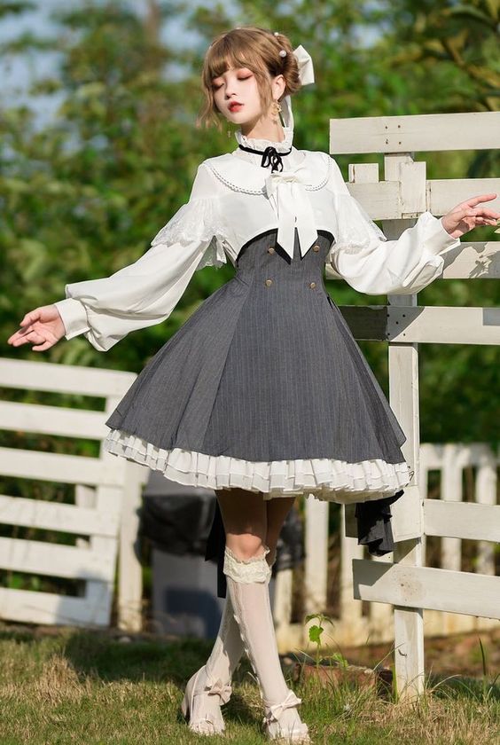 a young woman in a classic lolita dress poses in front of a white fence. the dress is grey and white, with a ruffled skirt and a white blouse with puffed sleeves.  she is also wearing white tights and white shoes. the background is a green garden.