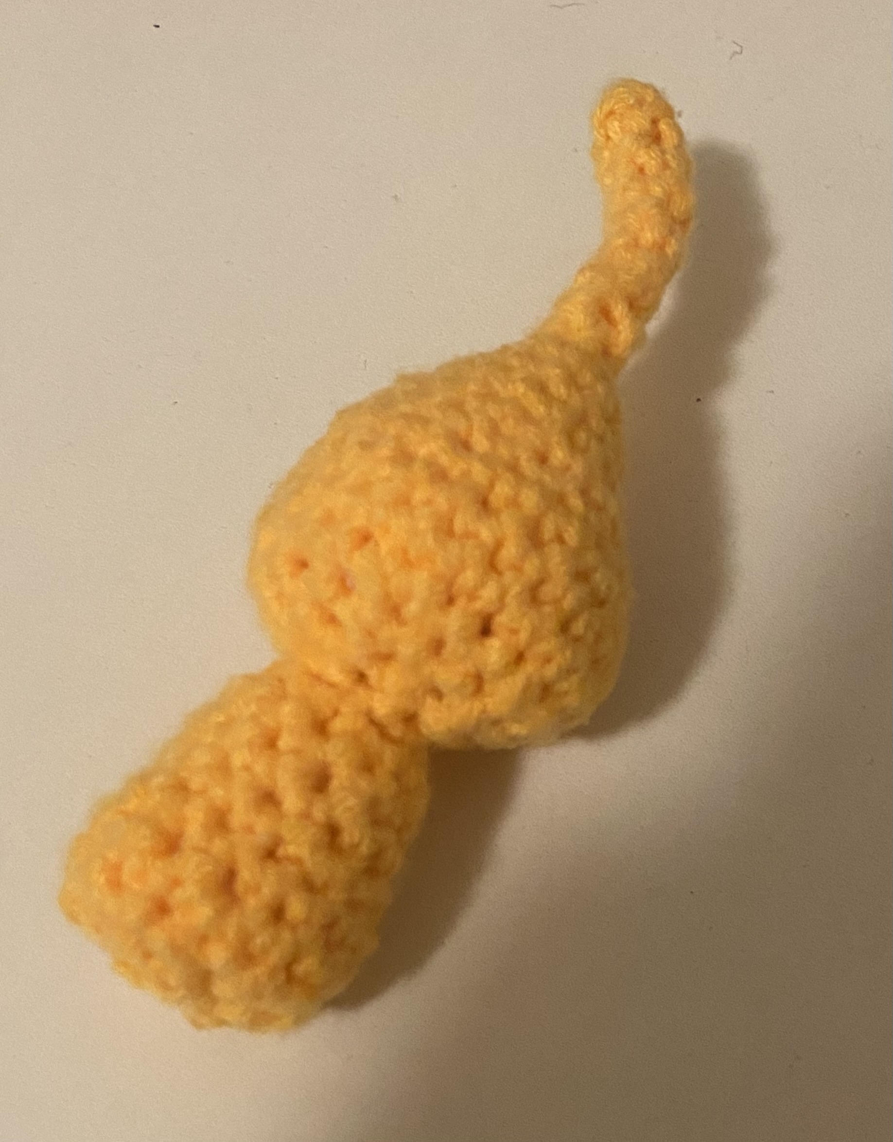 a crocheted yellow plush toy tail on a white surface. it appears to be made of yarn and has a bulbous shape with a curved end.