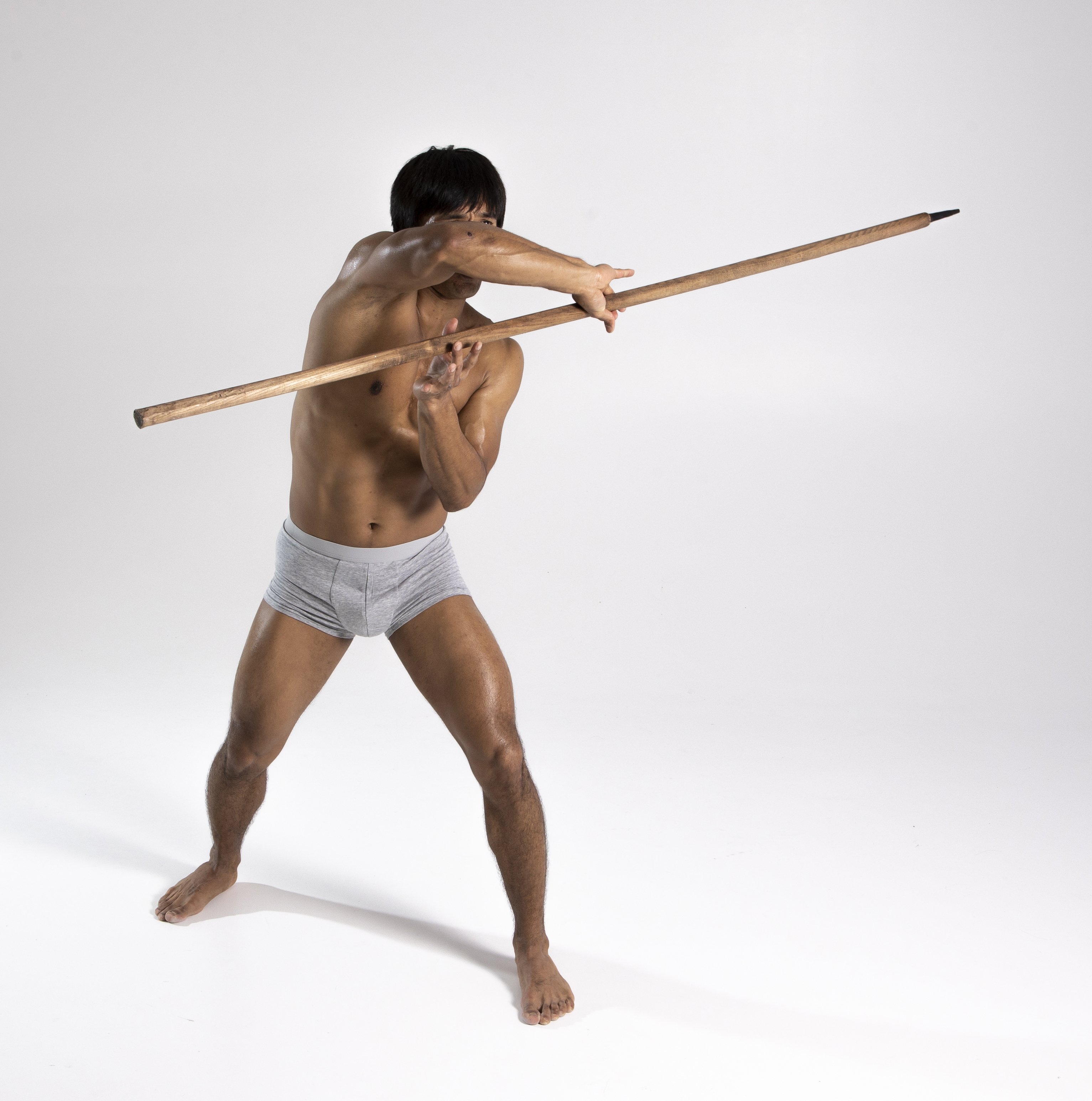 a shirtless, athletic man in grey underwear stands barefoot on a white background, holding a long wooden spear. he is in a defensive or attacking pose with his arms extended, gripping the spear in both hands. the studio shot highlights his physique and the spear's details.