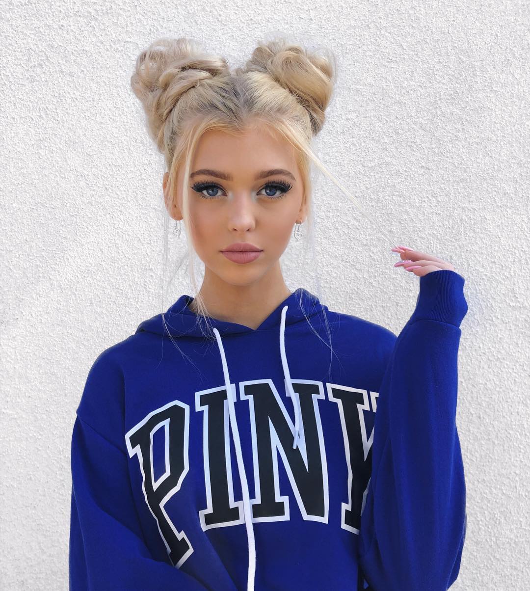 a portrait of a young woman with blonde hair styled in space buns. she's wearing a blue hoodie with "pink" printed in large white letters with black outline on the front. she is standing in front of a white wall, with her hand raised slightly. she has light makeup, prominent false eyelashes, and earrings. her fingernails are painted pink.