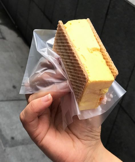 a hand is holding a yellow ice cream sandwich in a plastic bag.  the ice cream is between two waffle cookies and is a bright yellow color. the hand is holding the ice cream sandwich in front of a grey wall. the ice cream is melting slightly and the waffle cookies are starting to soften.  the person is enjoying a cold treat on a hot day.