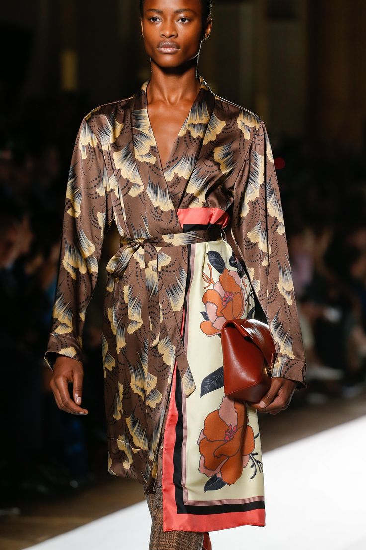 a model walks down a runway wearing a brown and beige silk robestyle dress with a large floral print. the dress is belted at the waist and features a contrasting orange and cream scarf detail at the hem.  the model carries a small brown leather handbag. the background is blurred but suggests a fashion show setting. the overall style is sophisticated and elegant.