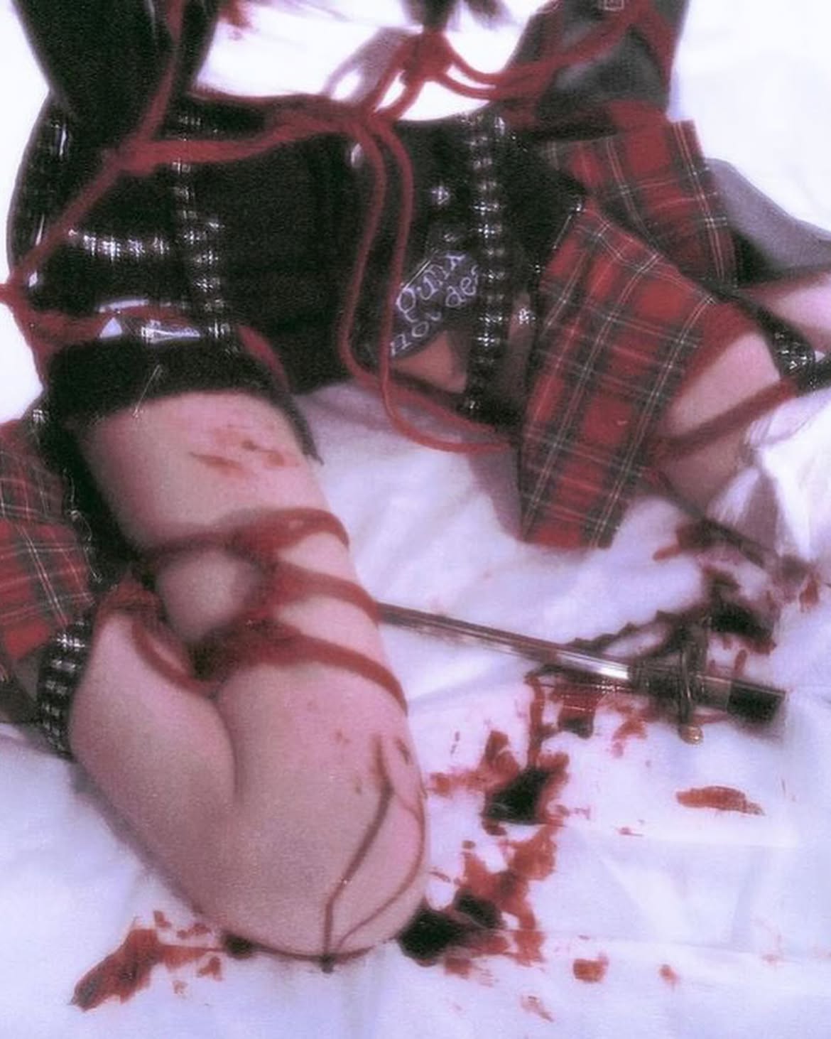 the image depicts a person in a black top and red plaid skirt, with their arm tied up and covered in blood. a blade lies nearby, suggesting selfharm. the scene is graphic and disturbing.