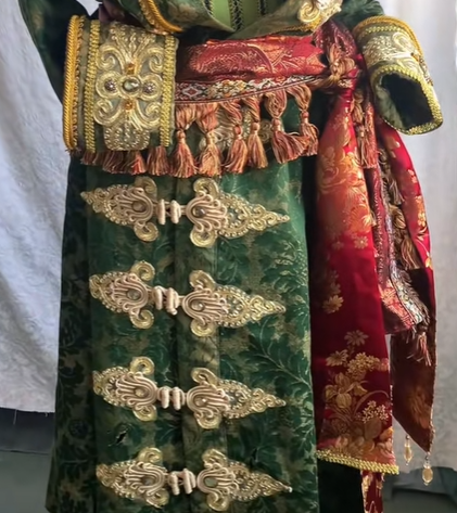 a detailed shot of a historical costume in shades of green, red, and gold. the costume features intricate gold trim, tassels, and ornate closures. it includes a red sash with gold detailing. the main fabric of the garment is green, with a pattern visible.