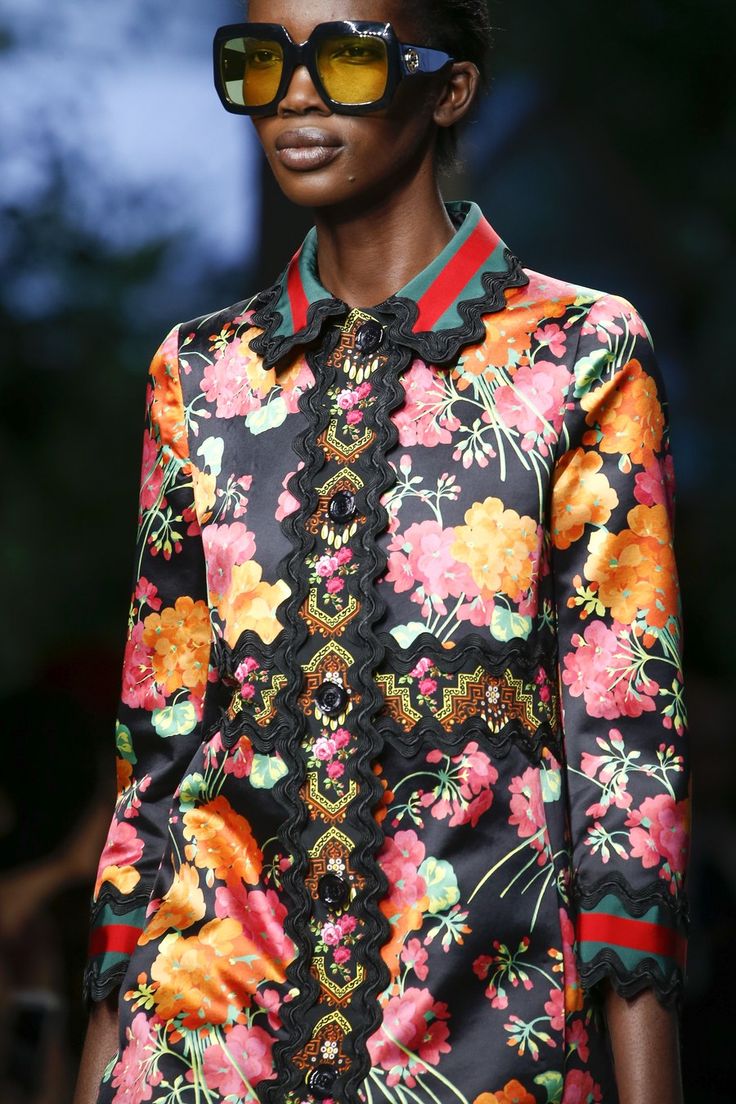 loseup shot of a female fashion model showcasing a gucci outfit on a runway. the model is a darkskinned woman wearing oversized yellowlensed sunglasses. her outfit is a black silk or satin coat with a vibrant floral print featuring oranges, pinks, and greens. the coat has intricate embroidery and detailing along the front placket and cuffs, which includes red and green accents. the overall aesthetic is one of high fashion, luxury, and elegance, typical of a gucci collection. the background is blurred, focusing the attention on the model and her attire.