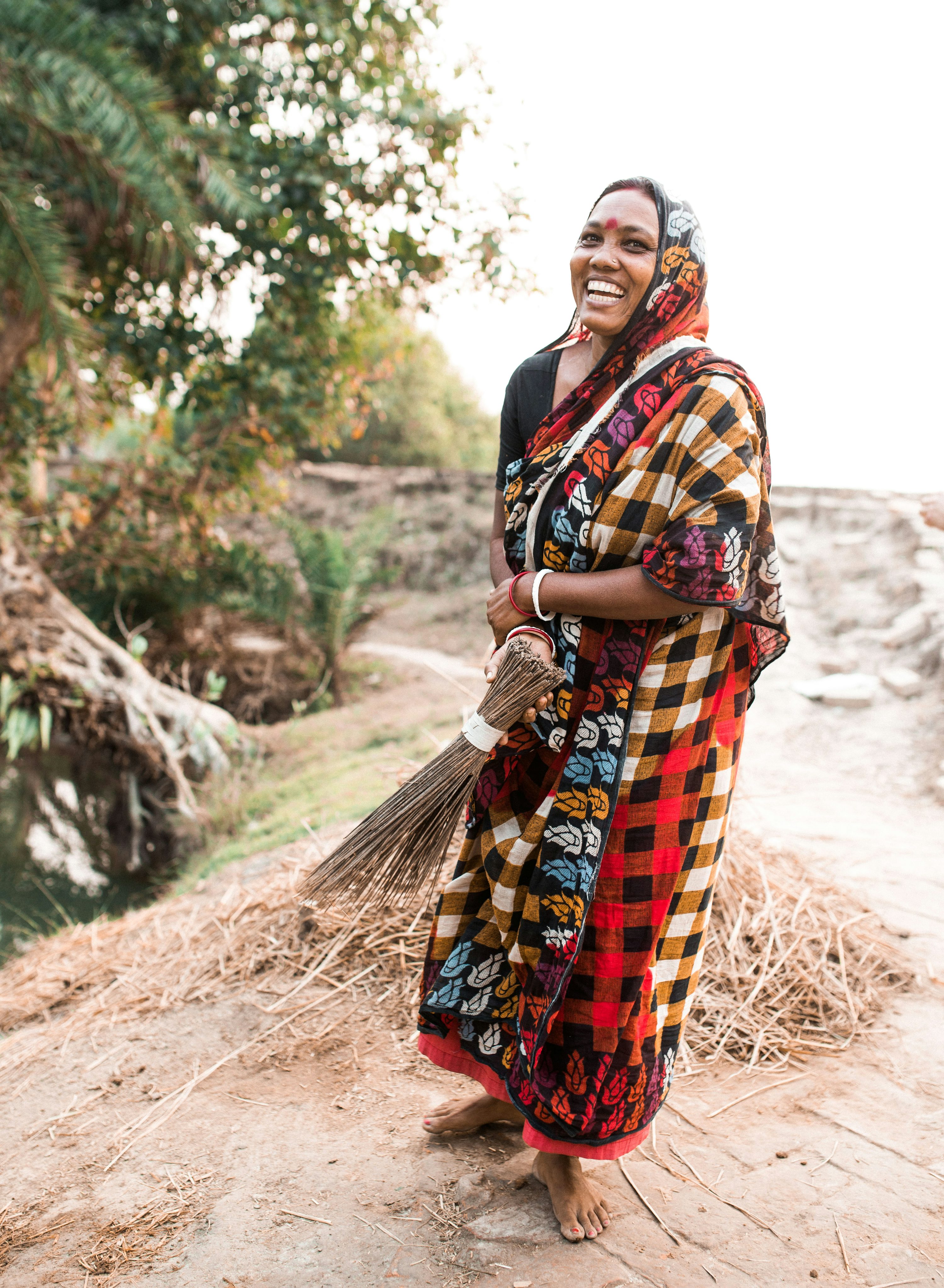 the image shows a smiling indian woman, dressed in a vibrant, colorful checked saree and a head covering. she holds a traditional broom made of natural materials. the setting appears to be a rural village in india, with dry straw and  rustic surroundings.  she looks happy and content, perhaps after completing some work.  the overall atmosphere is warm and positive. the photo captures a moment of everyday life in a rural indian setting.