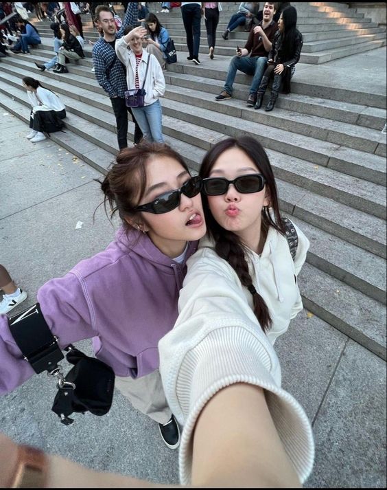 two young women pose for a selfie on a city street. they are standing on a set of wide stairs with other people walking and sitting in the background. the woman on the left wears a purple hoodie while the one on the right wears a white hoodie. both sport trendy sunglasses and smile playfully at the camera. the image has a casual and fun vibe with a wideangle perspective from the selfietaker. the overall color scheme leans towards cool grays and muted purples.