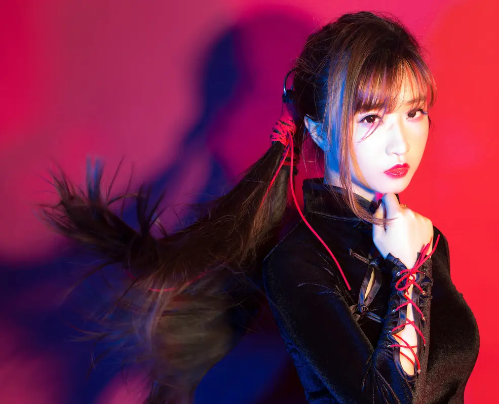 a woman with long black hair is posing in a red and black outfit, her hair is pulled back into a ponytail, her eyes are bright and she's wearing red lipstick. the background is a blurry red and blue.