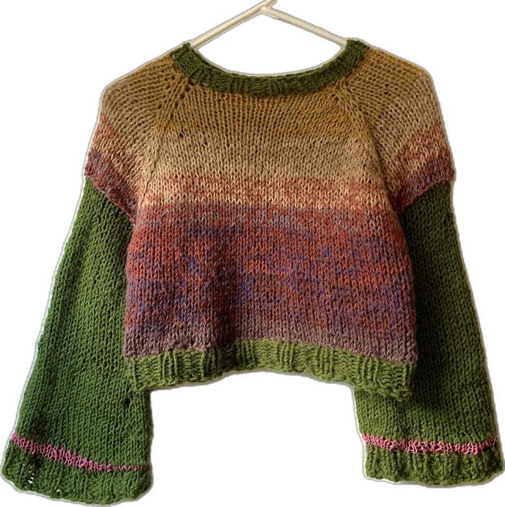 a closeup shot of a handknitted sweater. the sweater is in green, brown, and orange colors. it has bell sleeves and the trim is in pink. the sweater is laid flat on a surface.