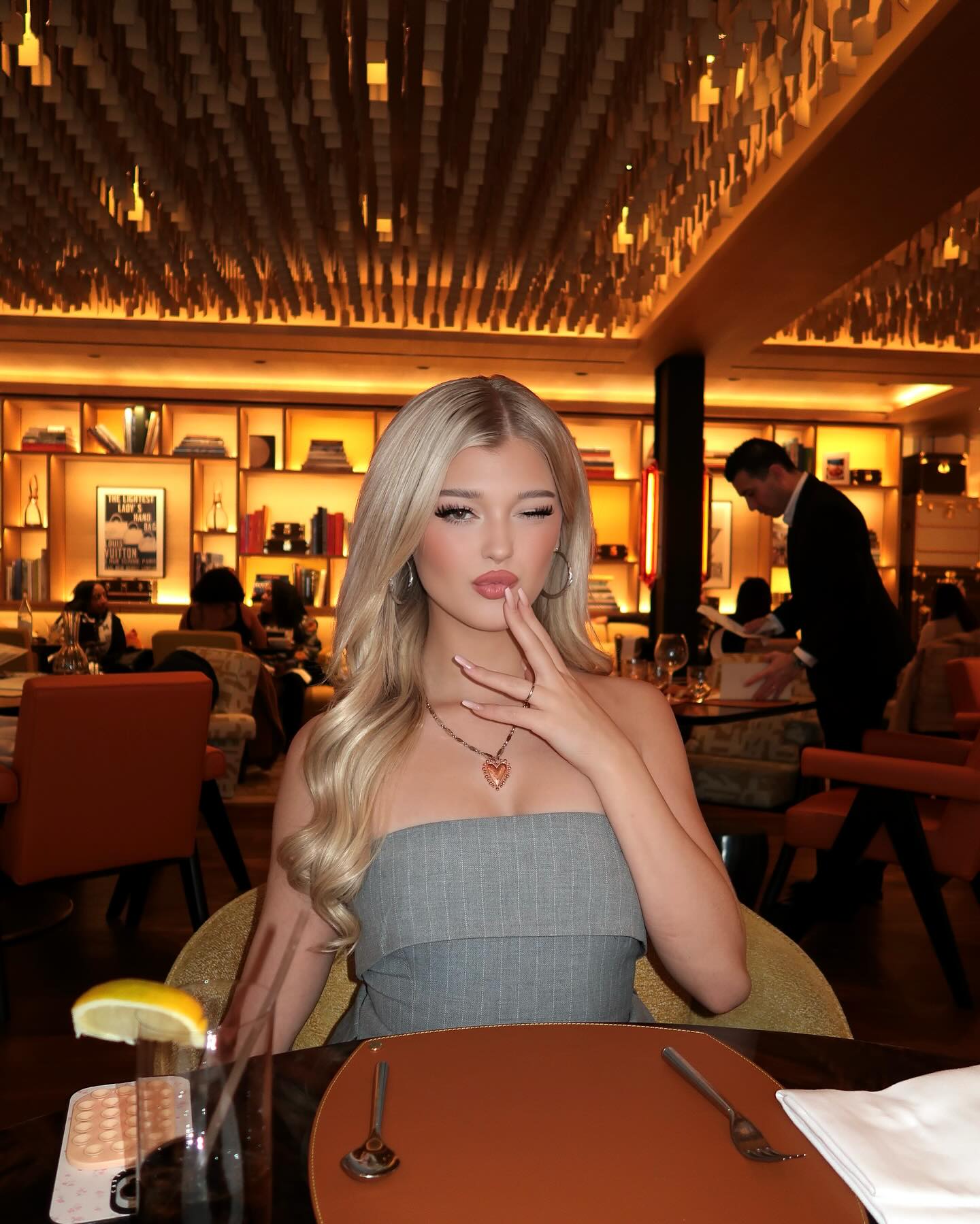 a young woman with blonde hair is sitting at a table in a restaurant. she is wearing a gray strapless top and a heart necklace. she is holding her hand to her mouth and pouting. there is a drink with a lemon slice on the table in front of her, as well as silverware and a napkin. a waiter is standing in the background.