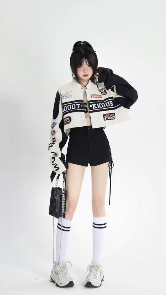 a young woman poses against a white background, showcasing a trendy streetwear outfit. she wears a cropped racing jacket with black and white panels and colorful patches, paired with a black crop top. her highwaisted black shorts feature tieup details on the sides. she completes the look with white sneakers, white socks, and a black chain handbag. her dark hair is pulled back, and she strikes a confident pose with one hand touching her hair.