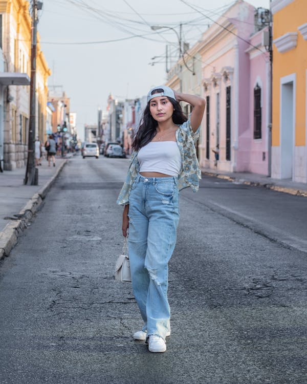 a young woman stands confidently on a city street, showcasing a casual yet stylish outfit. she's wearing light blue wideleg ripped jeans, a white crop top, a light blue patterned open shirt, and white sneakers. her accessories include a white baseball cap and a small white handbag.  the background features brightly colored colonialstyle buildings, suggesting a latin american setting, potentially mexico.  the overall vibe is relaxed and summery, capturing a candid portrait of the woman against the backdrop of a sunny street.