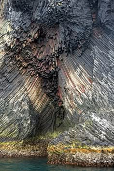 a closeup image of a cliff face with an intricate pattern of vertical rock formations, suggesting columnar jointing, possibly created by the cooling of basalt lava flows. the rocky texture is highlighted by the contrasting shades of grey, brown, and green. the cliff appears to be located near the sea, with the edge of the water visible at the bottom of the image. the overall effect is of a dramatic and rugged landscape, with a sense of power and geological wonder.