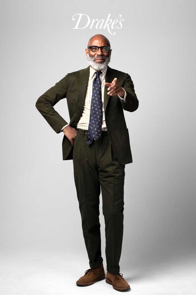 the image is a professional fashion photograph showcasing a man in a dark olive green tailored suit. he is an older man with gray hair and a beard, wearing glasses and brown suede derby shoes.  he's smiling and gesturing with one hand towards the viewer. the background is a simple, light gray.  the drake's brand logo is prominently displayed at the top. the overall impression is one of sophisticated, highend menswear.