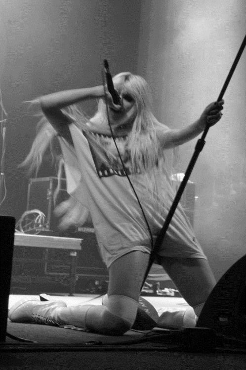 a black and white image shows a woman with long blonde hair kneeling on a stage while singing into a microphone. she is wearing a tshirt and knee pads. her long boots are visible. she is performing during a concert.