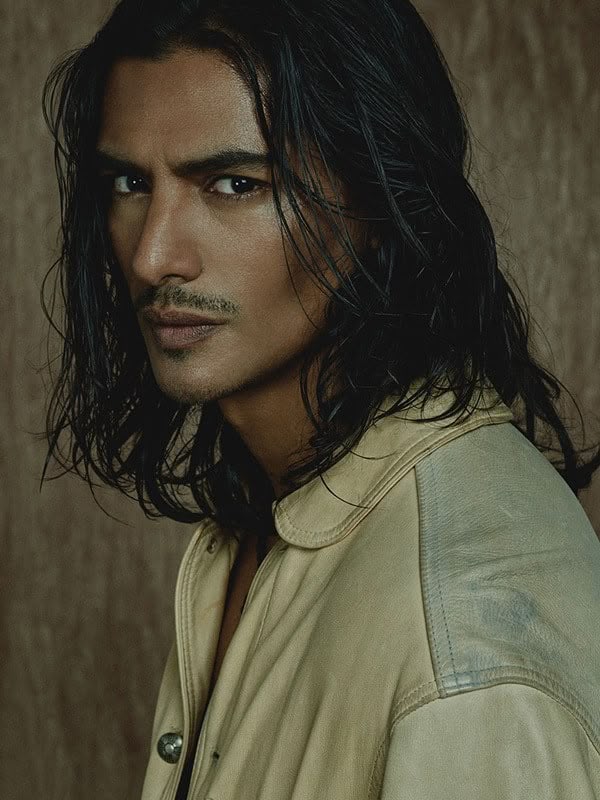 a portrait of a male model with long, dark hair, tanned skin, brown eyes, and a mustache. he has a serious expression and is wearing a tan leather jacket. the background is a neutral color.