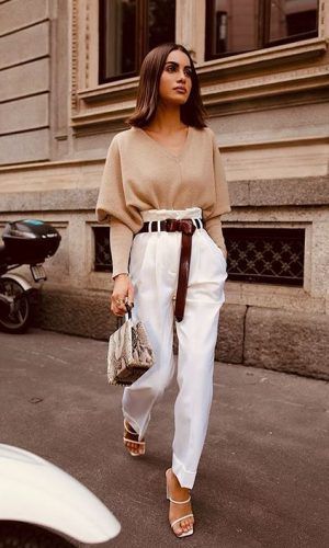 a stylish woman is captured walking down a city street. she's wearing a beige, oversized sweater, highwaisted white pants, a brown belt, and white heeled sandals. she carries a snakeskin purse. the overall style is casual yet chic and elegant. the backdrop is a city building. the image is welllit and showcases the woman's fashionable attire.