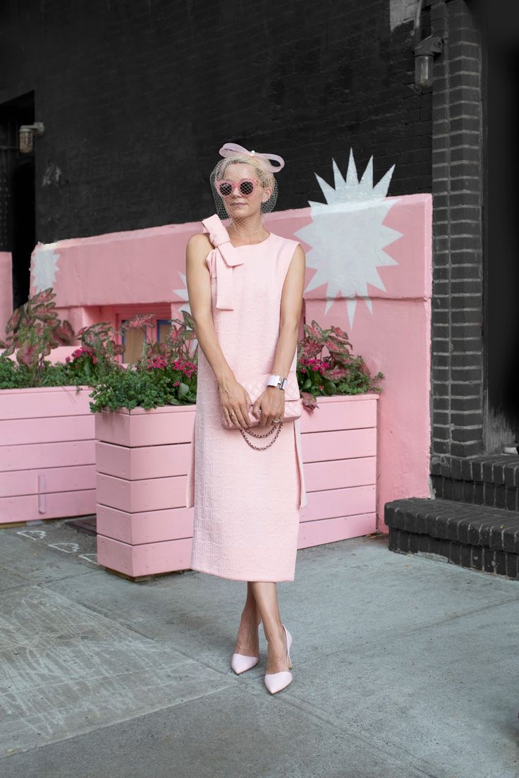 a stylish woman in a light pink dress and matching accessories stands on a city street in front of a pink wall with white starburst designs. she is wearing pink heels, a pink handbag, sunglasses and a pink hat with a veil. there are planter boxes with flowers next to the pink wall.