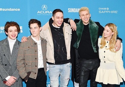 a group photo of five young actors at the sundance film festival.  from left to right: gaten matarazzo (in a grey coat), jacob bertrand (in a plaid jacket), machine gun kelly (in a beige coat),  and suki waterhouse (in a white sweater). noah jupe's face is partially obscured, but he's visible behind jacob bertrand. they are all smiling and posing for the camera against a blue background featuring various sponsor logos. the atmosphere is casual and celebratory, typical of a film festival premiere.