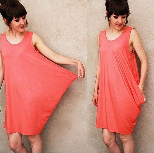 a woman is shown in a splitscreen image wearing a loose, coralcolored dress. on the left, she stretches out the dress's fabric with her hand, demonstrating its ample cut. on the right, she's shown with the dress hanging naturally, a pocket evident in the design. the background is a simple, textured wall.