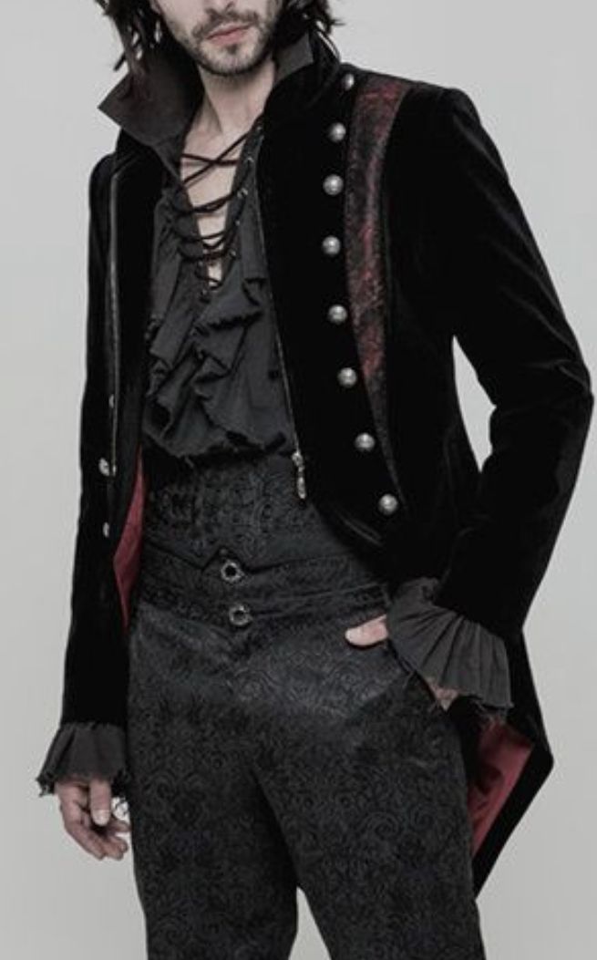 a man dressed in gothic victorian style stands against a light background. he wears a black velvet coat with a high collar, buttondown closure, red accents on the shoulder, and ruffled sleeves. underneath, he has a black laceup shirt with ruffles. the dress pants are black and patterned. he has a hand in his pocket.