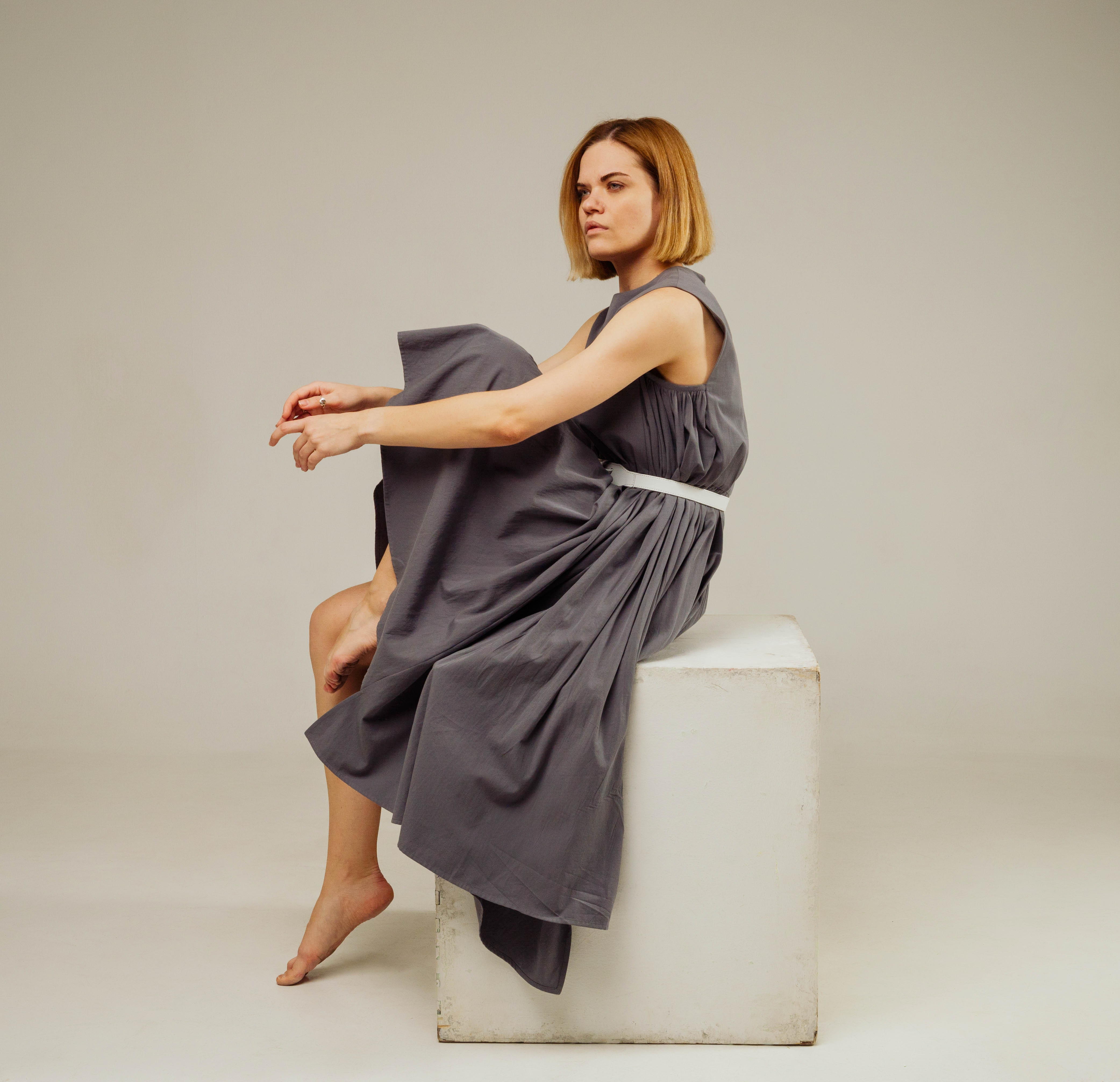 a woman in a long, gray dress sits on a white cube. she is looking off to her left, and her legs are crossed. the background is a light gray. the lighting is soft and the perspective is at eye level.