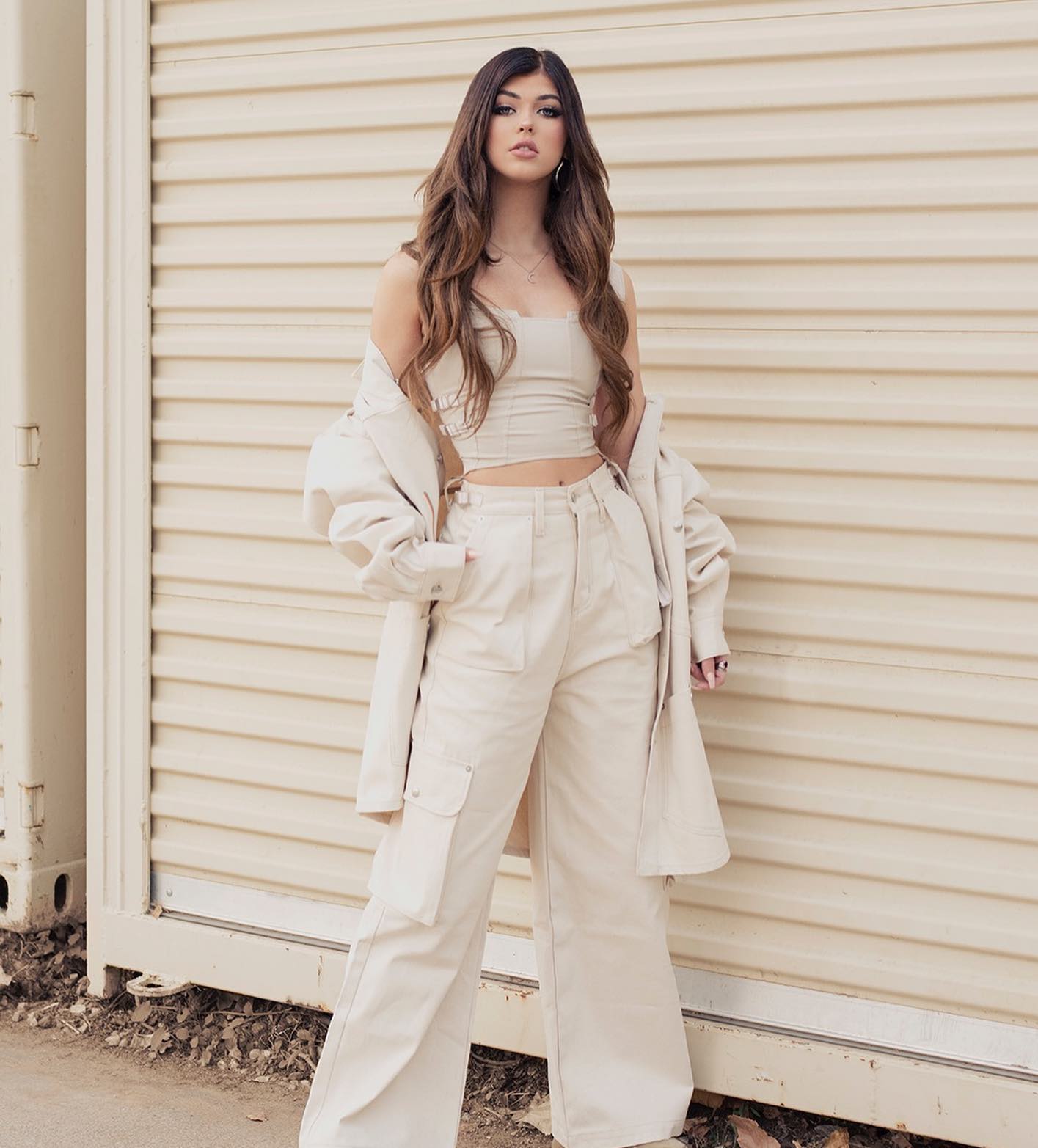 a woman stands facing forward in front of a beige wall, wearing beige clothing consisting of a top, pants, and jacket. she has long, brown hair.