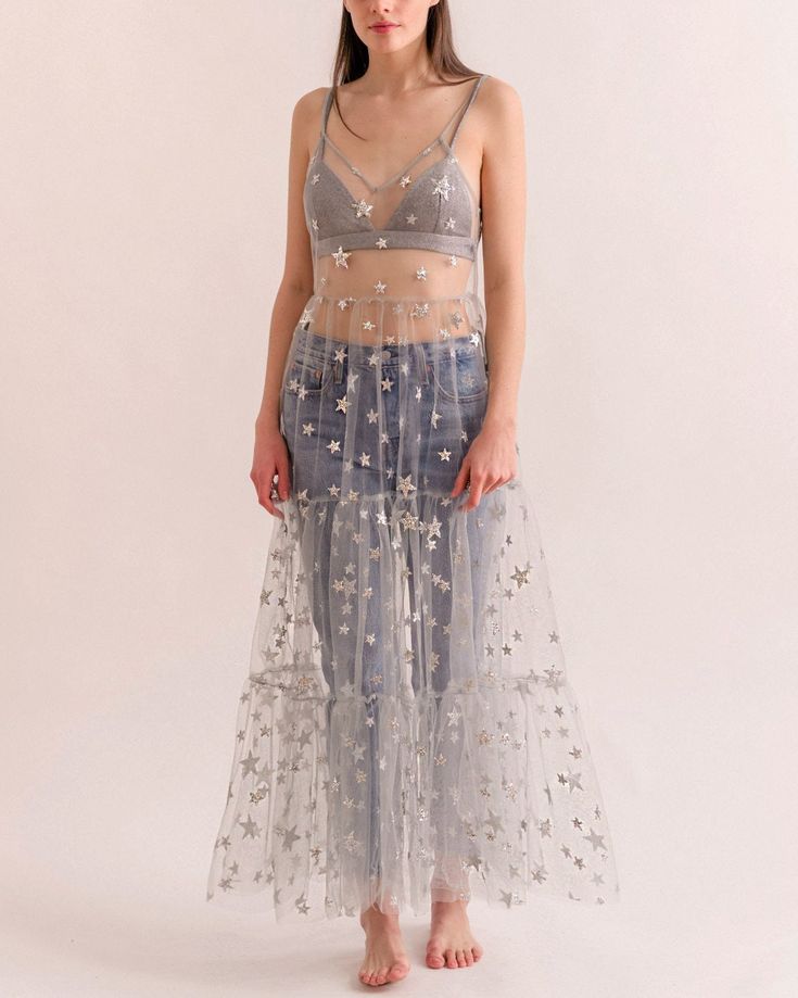 a woman in a long silver mesh dress with stars is posing in a studio. the dress is sheer and flows elegantly around her. she looks glamorous and elegant.