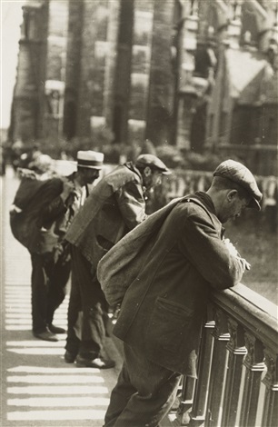 a poignant black and white photograph captures a group of men, seemingly unemployed or impoverished, standing on a bridge or elevated walkway. the men are dressed in simple, worn clothing, and some carry bags or packs suggesting they are transient.  they stand leaning against a decorative railing, their postures reflecting a sense of weariness or contemplation. the background features a blurred but visible backdrop of stone buildings, indicating an urban setting, possibly from the 1930s depression era. the overall mood is one of quiet contemplation and social commentary. the photograph's style is documentary realism, focusing on capturing a specific moment in time and conveying a sense of social reality.
