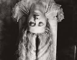 the image is a striking black and white portrait of a woman with long, flowing hair. she is positioned upside down, her head resting near the top of the frame, her body draped over what seems to be a lightcolored surface or piece of furniture. her expression is serene, almost melancholic, and her eyes are closed. she is wearing a lacetrimmed garment, and her hair is styled in a way that emphasizes its length and texture. the background is dark and shadowy, contributing to the overall dramatic and slightly surreal atmosphere of the photograph.  the overall style is reminiscent of classic portrait photography, possibly with influences from the art deco or film noir eras.