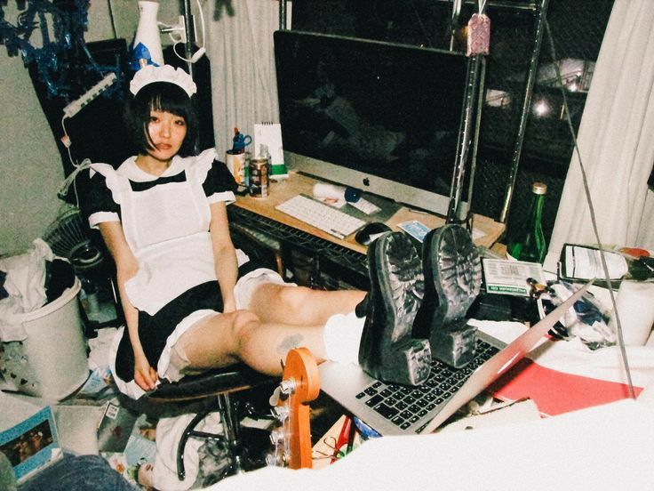 a woman dressed in a maid costume sits in a chair with her feet propped up on a laptop. the room is messy and cluttered with various items on the desk and floor, including a computer monitor, keyboard, a laundry basket filled with clothes, and a green bottle.