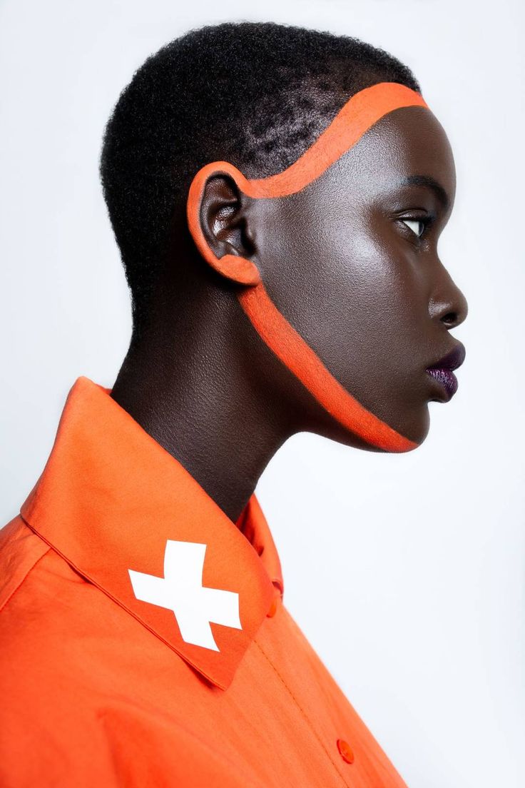 a close up profile portrait of a female model, she has short black hair, she is wearing a bright orange shirt with a white cross on the collar. she has orange paint lines on her face.  the backdrop is a white plain wall.