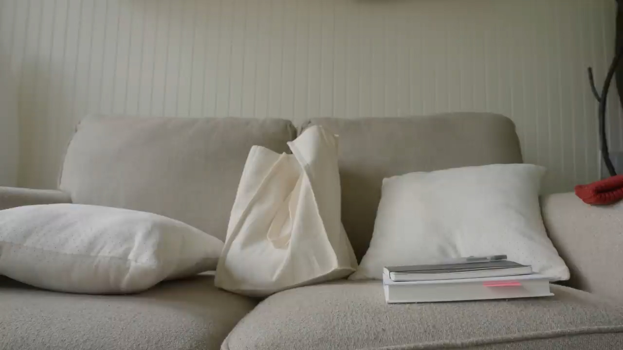 a beige couch with three white pillows, a white tote bag, a book and a notepad resting on it. the setting appears to be a living room bathed in soft, natural light.