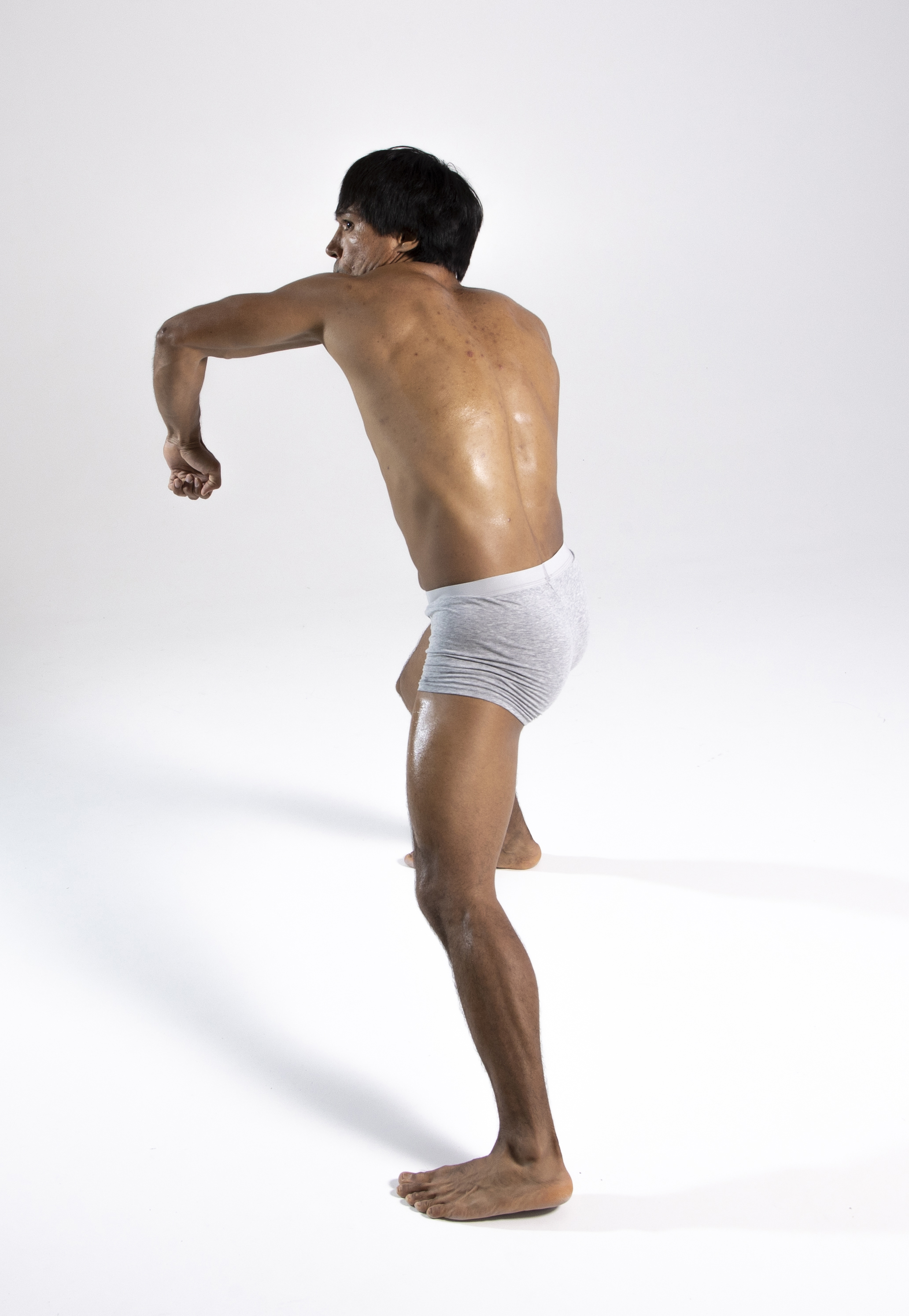 a barechested, muscular man is posing in a studio against a white background. he is wearing grey underwear and flexing his muscles. the view is from his back, showing his athletic build and healthy physique.