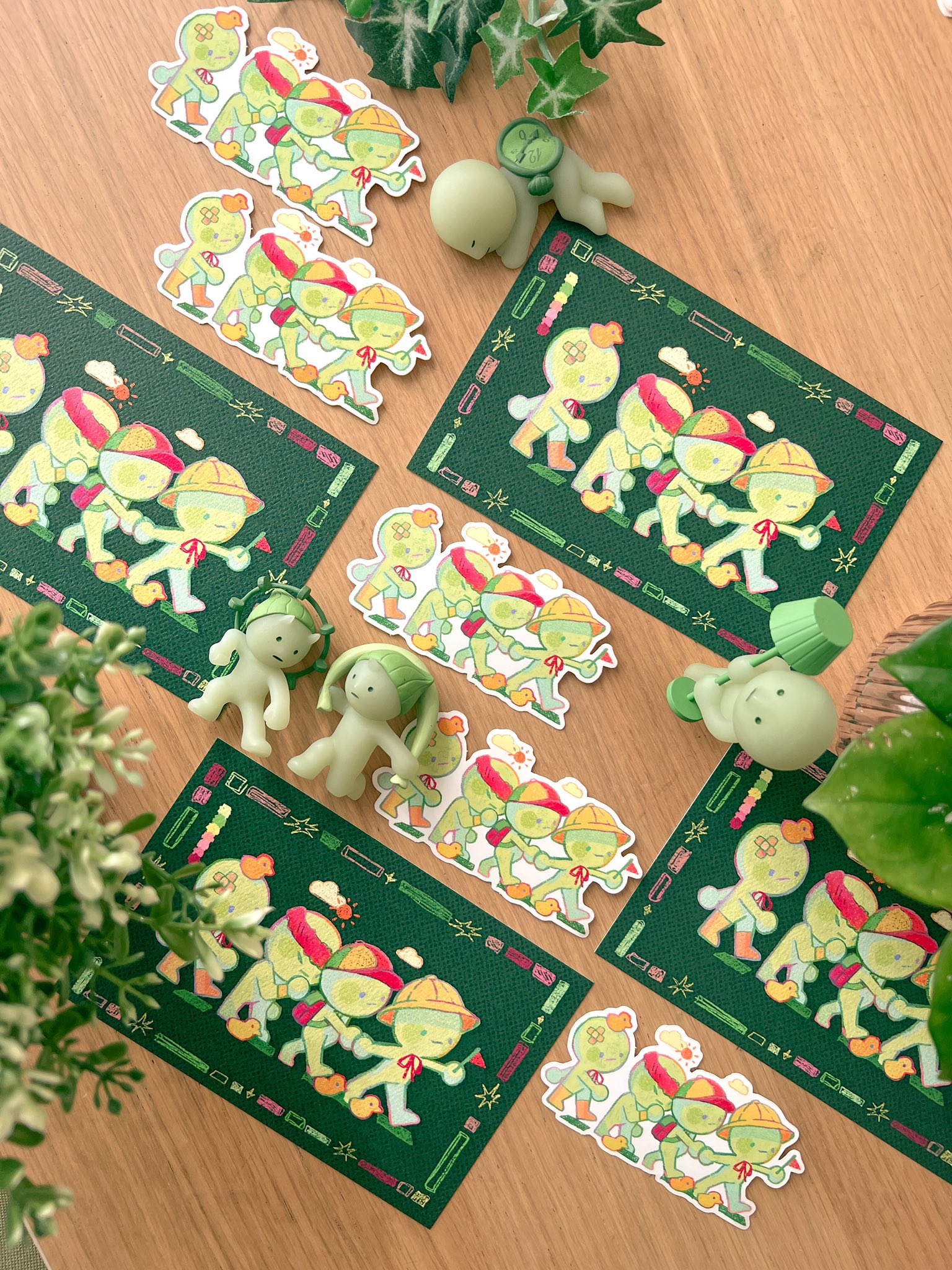 the image showcases a collection of charming green cartoon character stickers arranged on a light brown wooden surface. the stickers depict small, friendlylooking figures in various poses, possibly engaged in a group activity. several of the stickers are organized in sheets, and other individual stickers lie around.  small green plant figurines are also placed amongst the stickers, adding to the whimsical nature of the scene. the background includes a small potted plant and a wooden surface, hinting at a crafting or creative workspace. the overall style is cute, playful, and reminiscent of handmade goods or independent art.