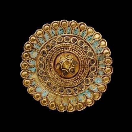 a closeup image of a round, golden ornament against a black background. the ornament is intricately decorated with concentric circles of small, raised dots and a central floral motif. the outer rim of the ornament features small, teardropshaped protrusions. the color palette of the ornament consists mainly of gold, with hints of turquoise blue in the background of the central design.
