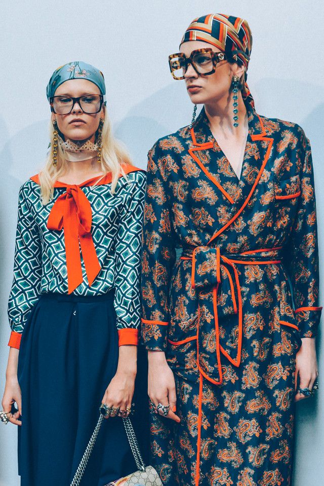 wo female models are showcasing stylish outfits. the model on the left is wearing a teal geometric print blouse with an orange bow, a dark blue aline skirt, and oversized eyeglasses.  she has a light blue headscarf and several rings and long earrings. she also carries a gucci handbag. the model on the right is wearing a dark blue paisley print silk robe with orange trim, a matching head scarf, oversized glasses, and long earrings.  both models display a coordinated and fashionable aesthetic. the background is a simple light gray wall. the image suggests a fashion shoot or runway show.