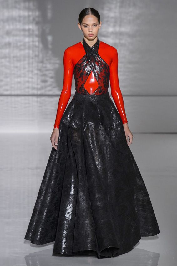 a woman is walking on a runway wearing a red and black gown. the dress has long red sleeves and a black bodice with a crisscross design at the neckline. there is a diamond shape cut out on the front of the bodice. the skirt of the dress is full and black, made of a shiny, textured fabric. the woman is tall and slender, and her hair is pulled back. the background is a blurry gray, suggesting a stage setting.