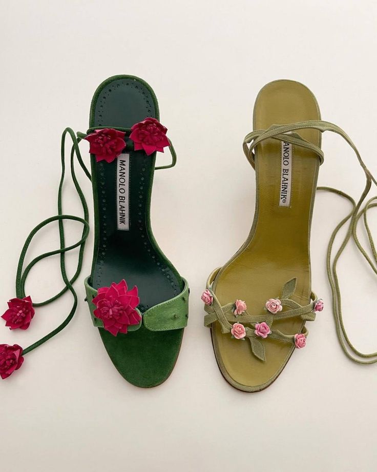 two manolo blahnik sandals are displayed sidebyside on a neutral surface. the left sandal is a deep green suede with large, bright pink floral embellishments on the strap and around the ankle tie. the right sandal is a muted olive green suede with smaller, light pink roselike floral embellishments along the strap. both sandals feature tie straps that wrap around the ankle and bear the manolo blahnik label on the insole.