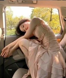 a young woman in a long, flowing dress is seated in the backseat of a car. she is leaning against the window, with her head resting on her arms. the car appears to be parked in a wooded area, and the sunlight is streaming in through the windows, creating a warm and inviting atmosphere.